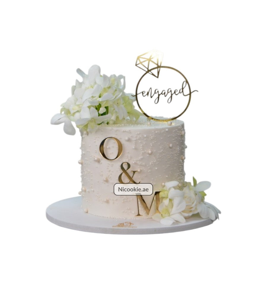 Elegant engagement cake with pearl details, white flowers, and a gold "engaged" topper.