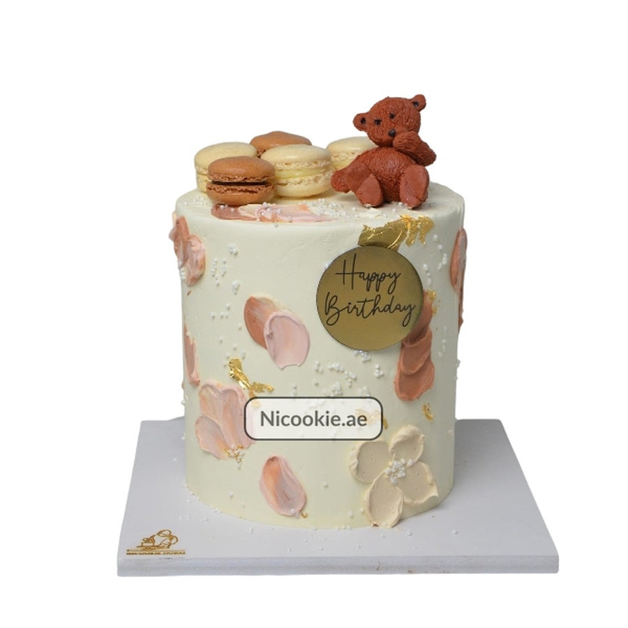 A white cake with a teddy bear, macarons, and floral accents.