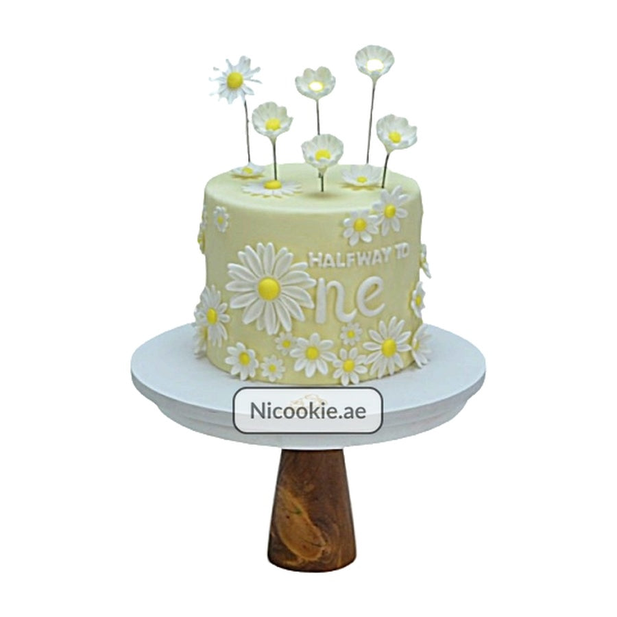 A yellow cake decorated with white daisies and the words "Halfway to One".