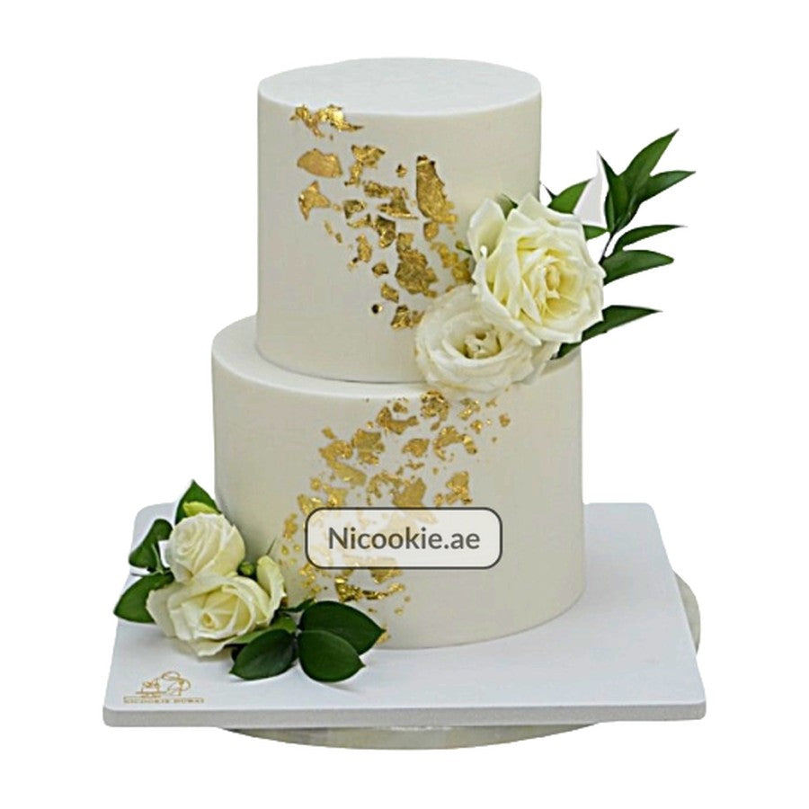 A two-tier white cake decorated with gold leaf and white roses.
