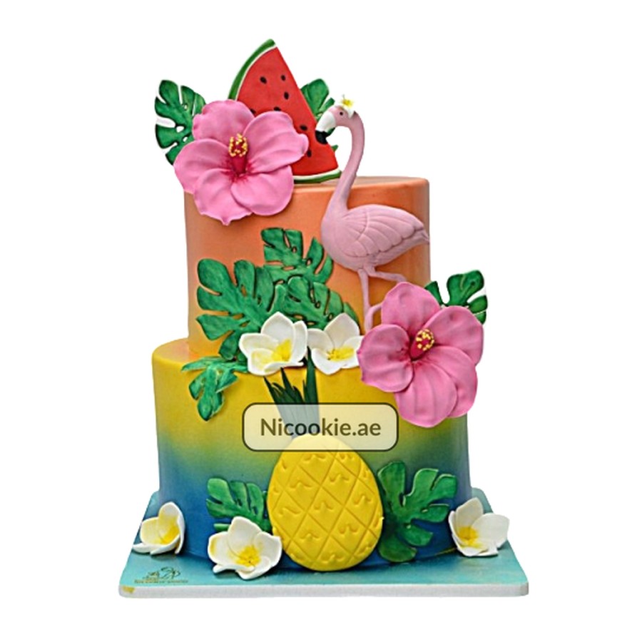 Two-tiered tropical cake with flamingo, hibiscus, pineapple, and watermelon decorations.