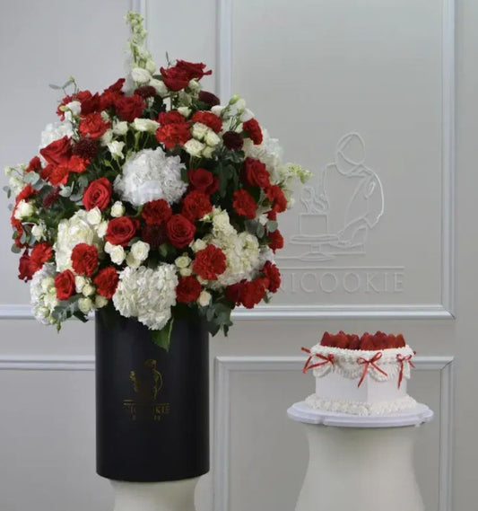 Red and white roses and hydrangeas in a black box with a white cake topped with strawberries.