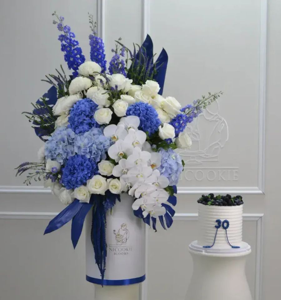 A blue and white floral arrangement with white orchids and hydrangeas sits next to a small white cake topped with blueberries.