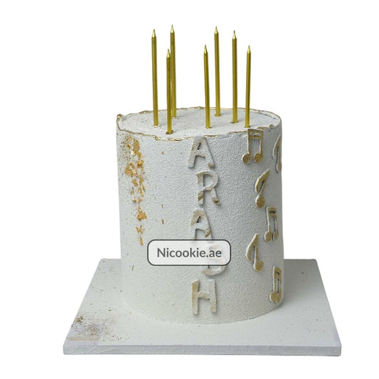 A white cake with gold musical notes and the name Arash, topped with gold candles.