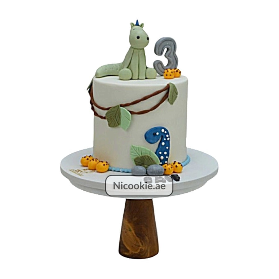 A white cake decorated with a green dinosaur, the number 3, and jungle elements.