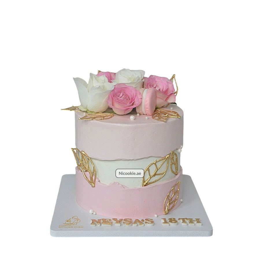 A two-tiered pink and white cake decorated with roses, macarons, and gold leaves.