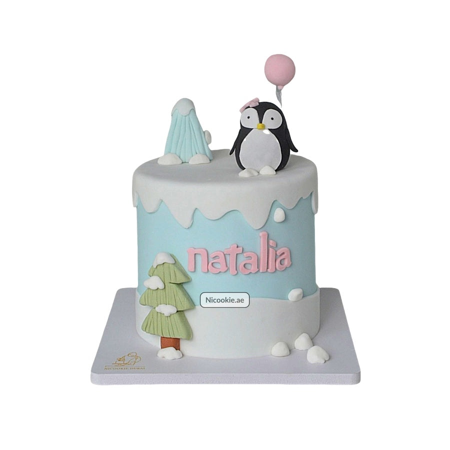 A winter-themed cake with a penguin, balloon, tree, and the name Natalia.