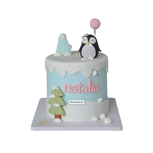 A winter-themed cake with a penguin, balloon, tree, and the name Natalia.
