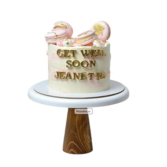 A white cake with pink accents, gold lettering, and pink macarons on a wooden cake stand.
