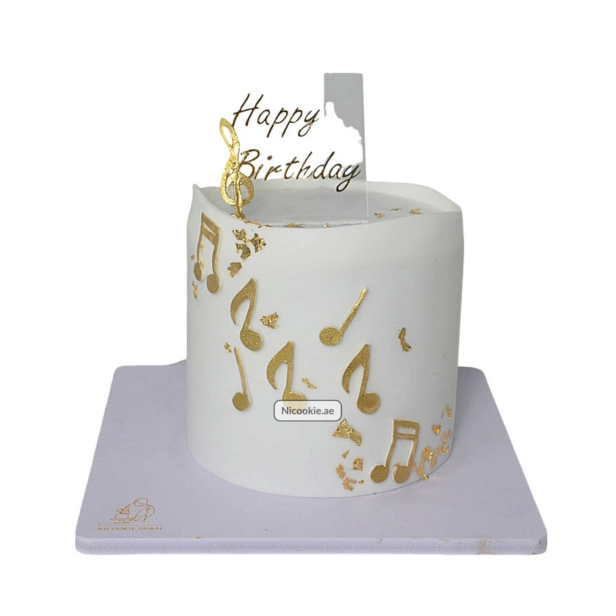A white cake decorated with gold musical notes and a "Happy Birthday" topper.