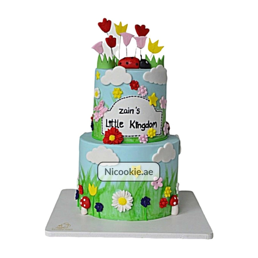 A two-tiered cake decorated with a garden theme, flowers, ladybugs, and clouds.