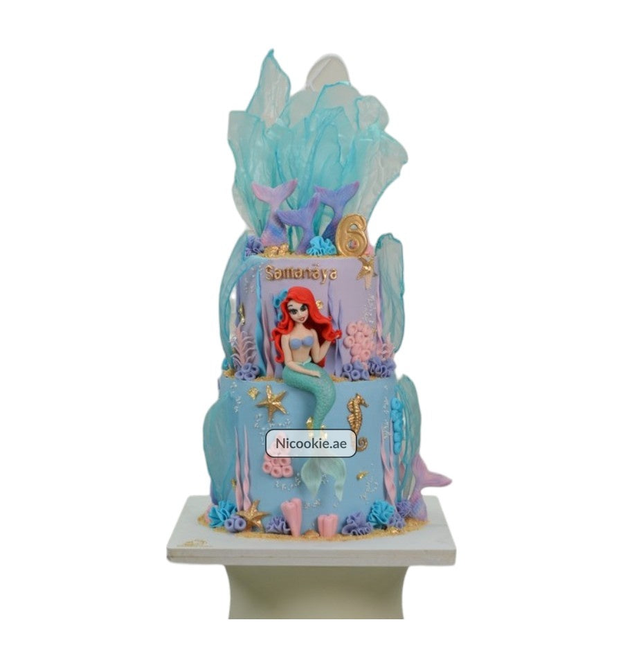 A two-tier mermaid-themed cake with Ariel, mermaid tails, and ocean decorations.