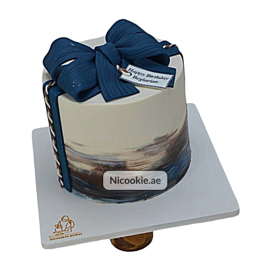 A round cake decorated with blue and brown brushstrokes, topped with a navy blue bow and a birthday tag.