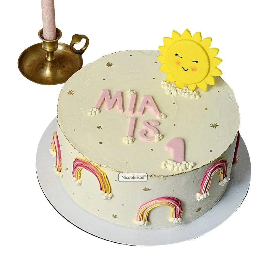 A first birthday cake with a sun, rainbows, and "Mia is 1" text.