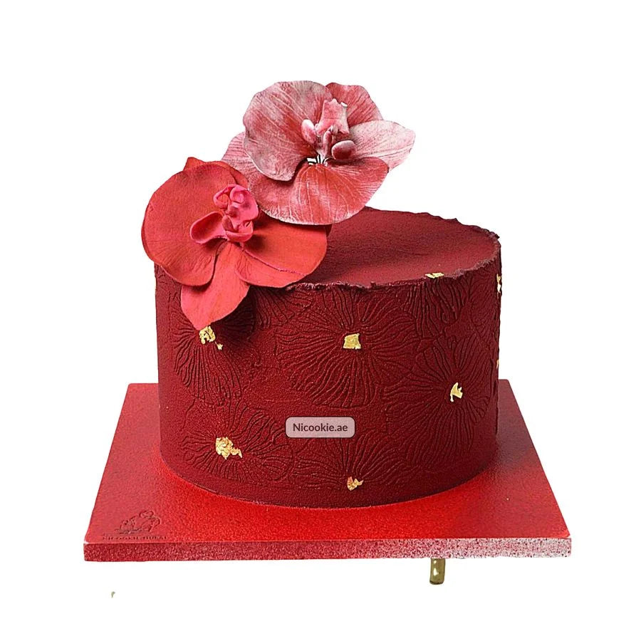 A red cake with embossed floral patterns and gold leaf accents, topped with two red sugar orchids.