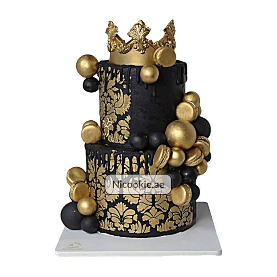 A two-tiered black cake with gold floral patterns, macarons, and spheres, topped with a gold crown.