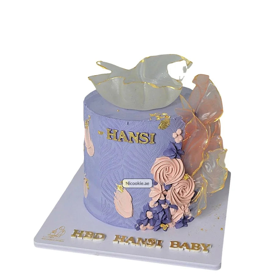 A lavender cake with gold lettering, flowers, and isomalt sails.